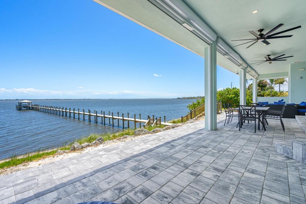 A spacious patio offers a relaxing area overlooking the water, featuring a dining set and comfortable seating. A long wooden dock extends into the river, with clear blue skies visible in the background. The outdoor space is paved with large stone tiles, enhancing the serene atmosphere.