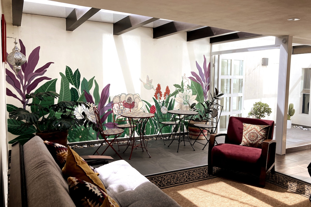 A cozy communal area features a mural of plants and flowers enhancing the atmosphere. Two round tables with metal chairs are positioned beside a gray sofa. A large red armchair adds a pop of color, while natural light floods the space through large windows.