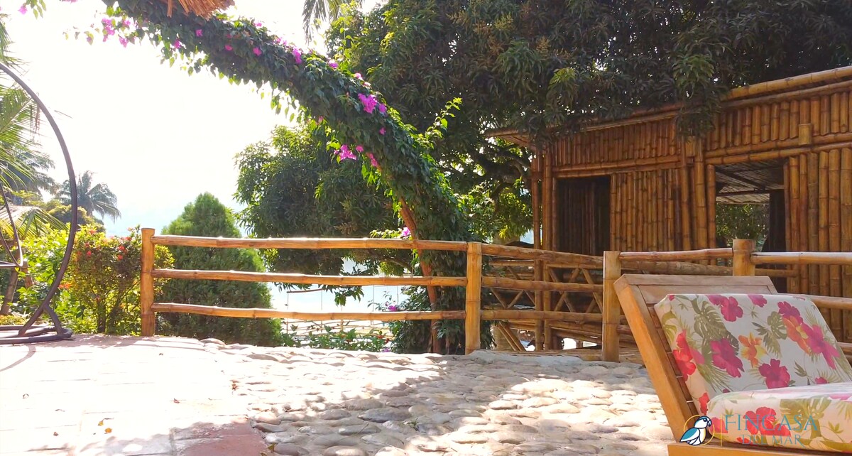 A tranquil outdoor seating area features a wooden fence and a comfortable lounge chair adorned with floral patterns. A lush green tree with vibrant purple flowers extends overhead, while hints of water can be seen in the background, creating a serene environment.