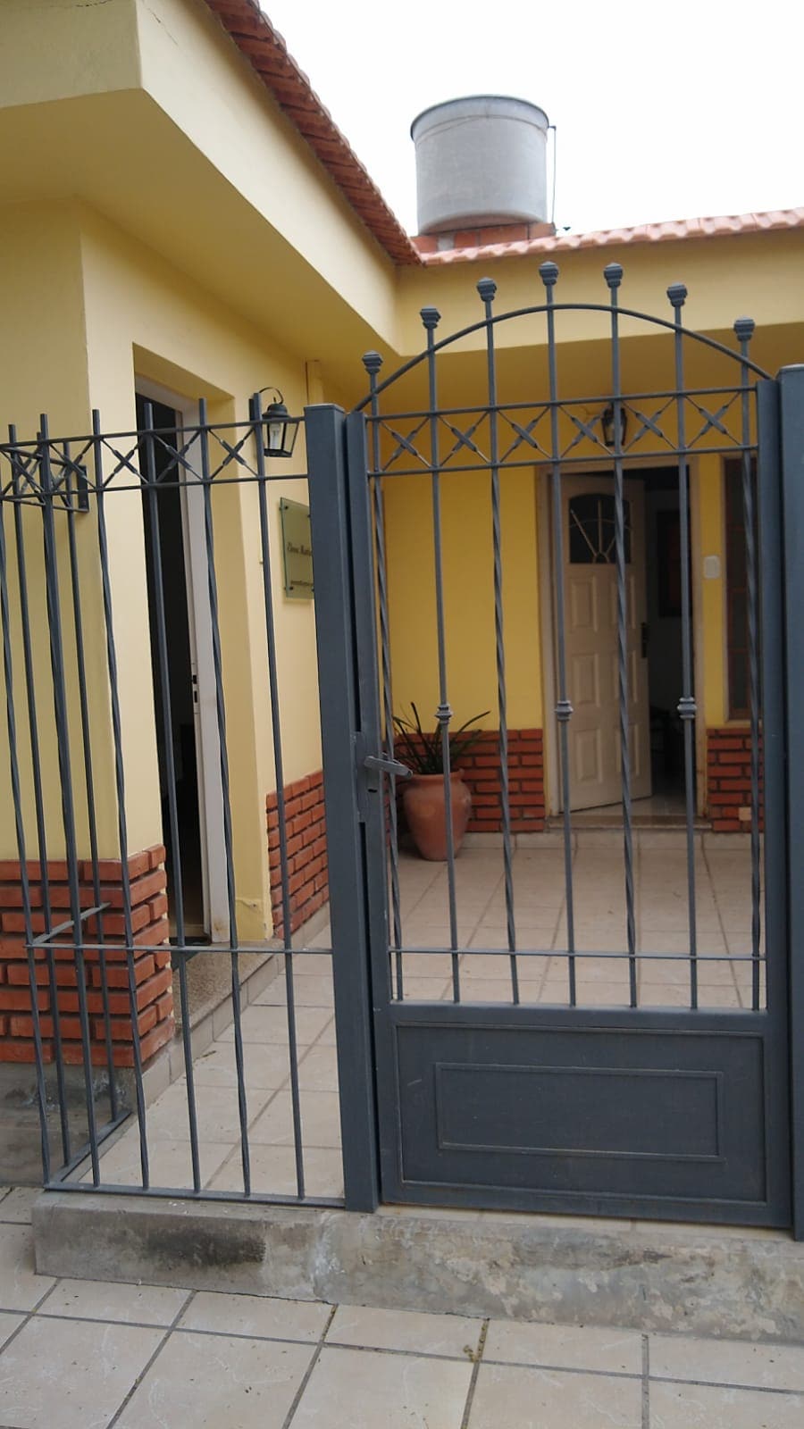 A gated entrance leads to a vibrant yellow house, showcasing a secure iron fence with a lockable door. The entrance features tiled flooring and potted plants that add a touch of greenery to the welcoming facade.
