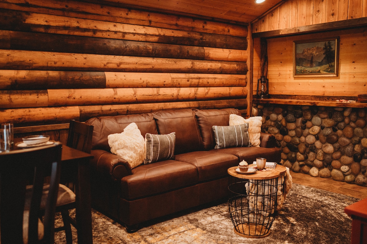 A cozy living area features a brown leather sofa adorned with decorative pillows. A round coffee table with light refreshments is set in front. The rustic log walls and stone accent provide warmth, while soft lighting enhances the welcoming ambiance.