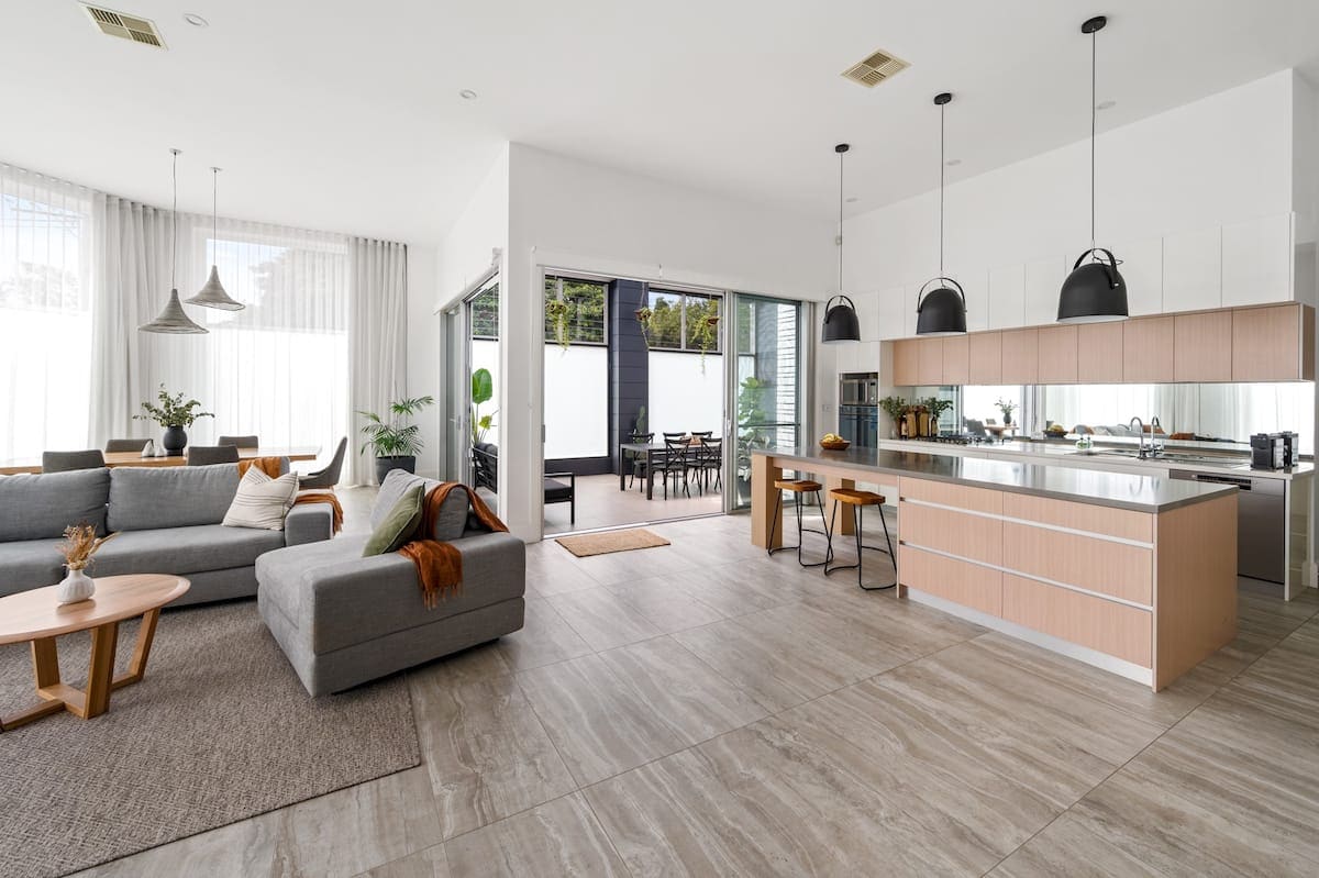 An open plan living area features a spacious kitchen with a large island, dining seating, and modern appliances. Natural light floods the room through large windows, illuminating a comfortable lounge space with a gray couch and stylish coffee table, creating an inviting atmosphere for relaxation and socializing.