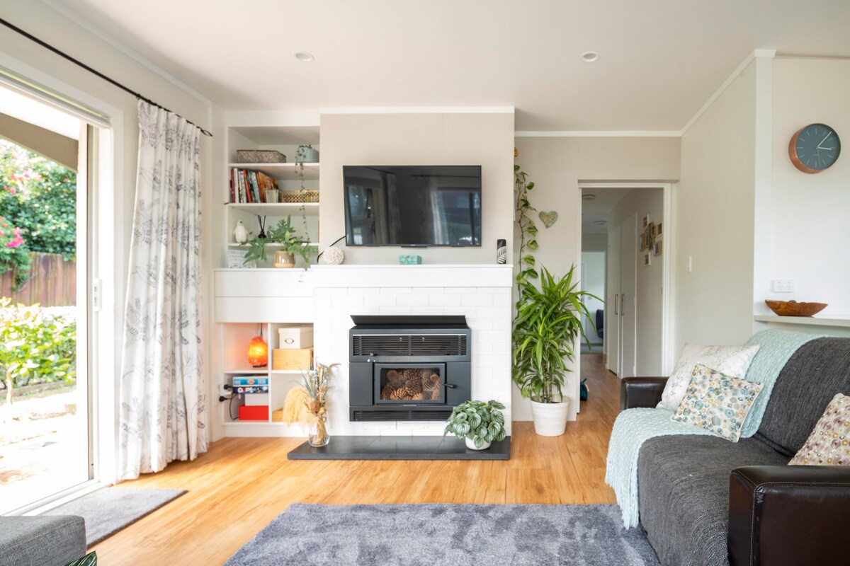 A living area features a cozy fireplace built into a white brick wall, complemented by a mounted TV above it. A bookshelf displays decorative items, while a comfortable gray sofa is positioned in front. Natural light enters through sliding glass doors that lead to the garden.