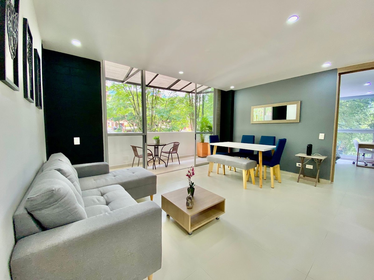 A spacious living area is equipped with a light gray sofa and a wooden coffee table, complemented by a six-seat dining table. Large windows provide natural light and views of greenery. A small table with two chairs is positioned on the terrace, enhancing the open layout.