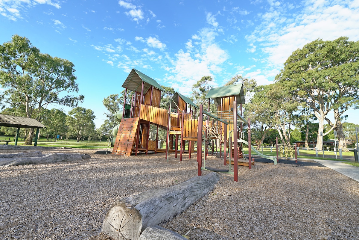 A vibrant playground structure is set against a clear sky, featuring multi-level play areas with slides and climbing features. Surrounding the playground, natural wood chips and logs create a safe play surface, complemented by lush greenery and open space in the background.
