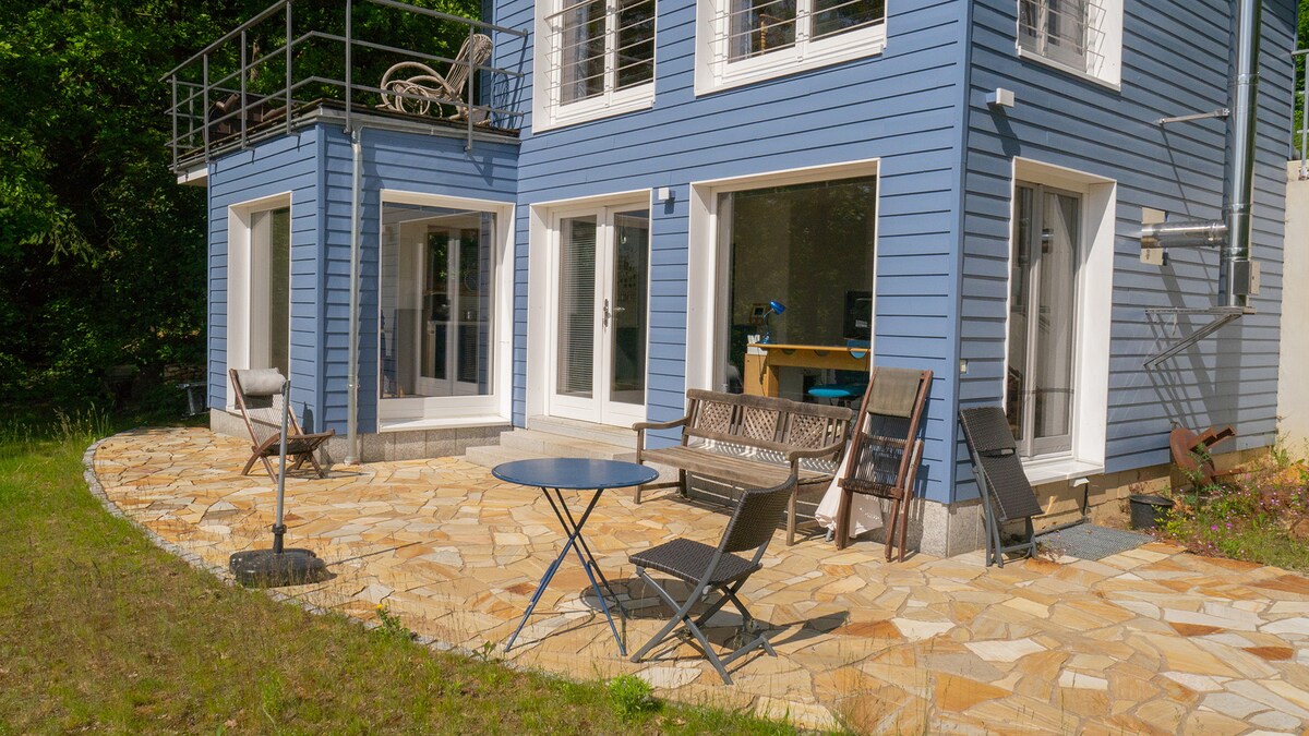 A light blue house features large windows and a modern design, surrounded by a stone patio. Outdoor seating includes a small round table and several chairs, providing a comfortable area for relaxation. Lush greenery frames the property, enhancing the tranquil environment.