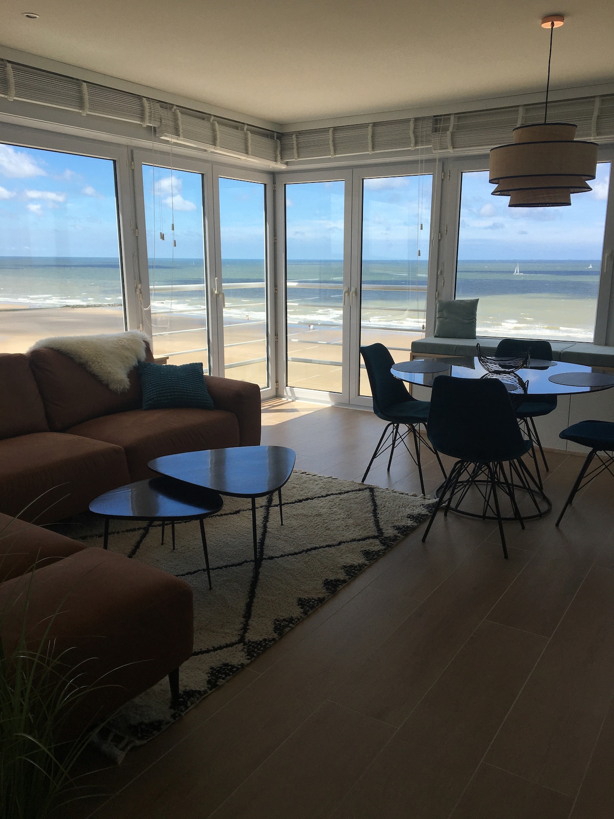 The living room features large glass doors that provide a direct view of the beach and ocean. A comfortable sofa is paired with small side tables, while a dining area with modern chairs and a round table is also visible. Natural light floods the space.