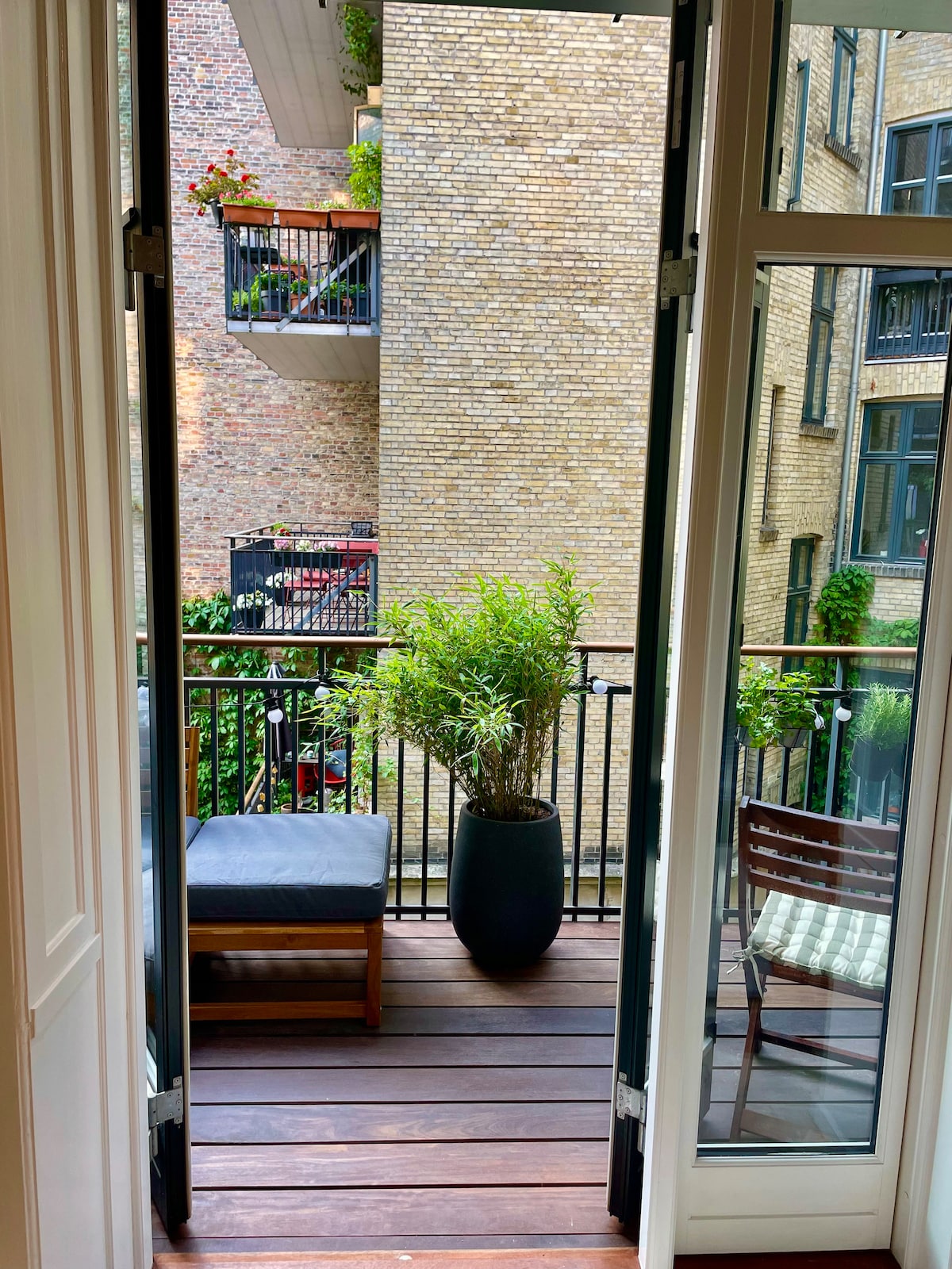 A balcony view is showcased, featuring a planter with greenery and a seating area that includes a lounge chair and wooden chairs. The space is framed by open double doors, with brick walls of neighboring buildings in the background.
