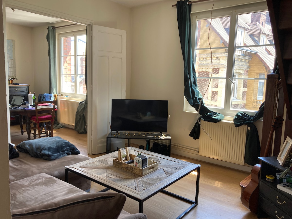 A living area is presented, featuring a comfortable sofa and a wooden coffee table adorned with decorative items. Two large windows allow natural light to illuminate the space, and a television is positioned against the wall. A dining area is visible in the background.