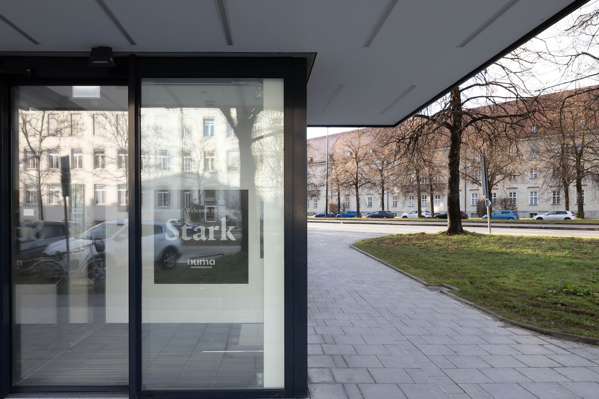 The exterior view displays a modern building entrance with large glass doors marked with the name 'Stark.' A tree-lined street is visible through the glass, alongside a path leading to the entrance and parked cars on the adjacent street.