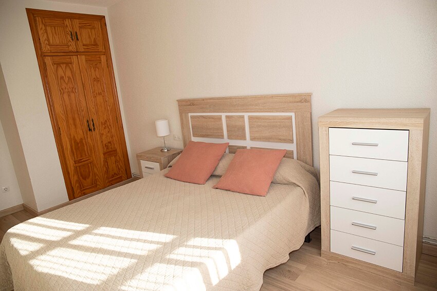 A well-lit bedroom features a spacious bed with a neutral-toned quilt and two coordinating cushions. A nightstand with a lamp is positioned beside the bed, while a modern dresser is placed against the wall. Natural light filters through a nearby window, highlighting the wooden closet.