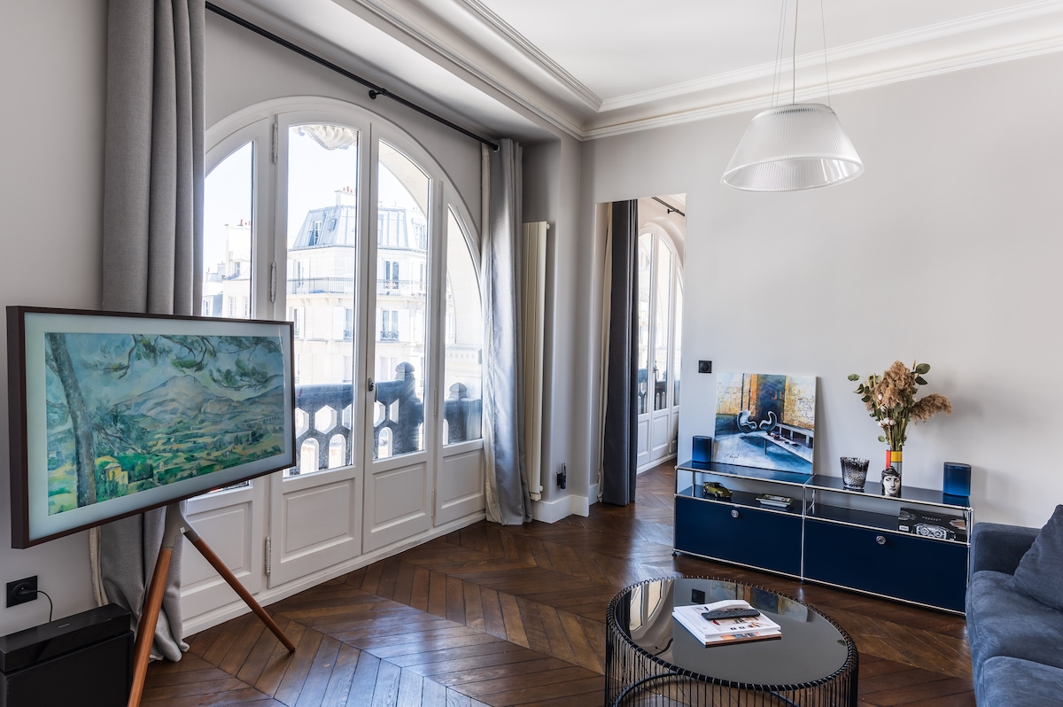 A bright living area features large arched windows allowing natural light to fill the space. A comfortable sofa is positioned near a sleek entertainment unit, which holds decorative items and a modern television. Artwork is displayed on an easel, adding color to the room.