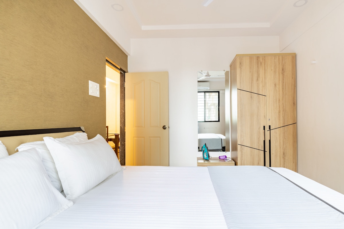 A bedroom is presented featuring a spacious bed adorned with crisp white linens. A light brown accent wall adds warmth, while a closet and dressing table are included in the room. Natural light enters through a nearby window, enhancing the inviting space.