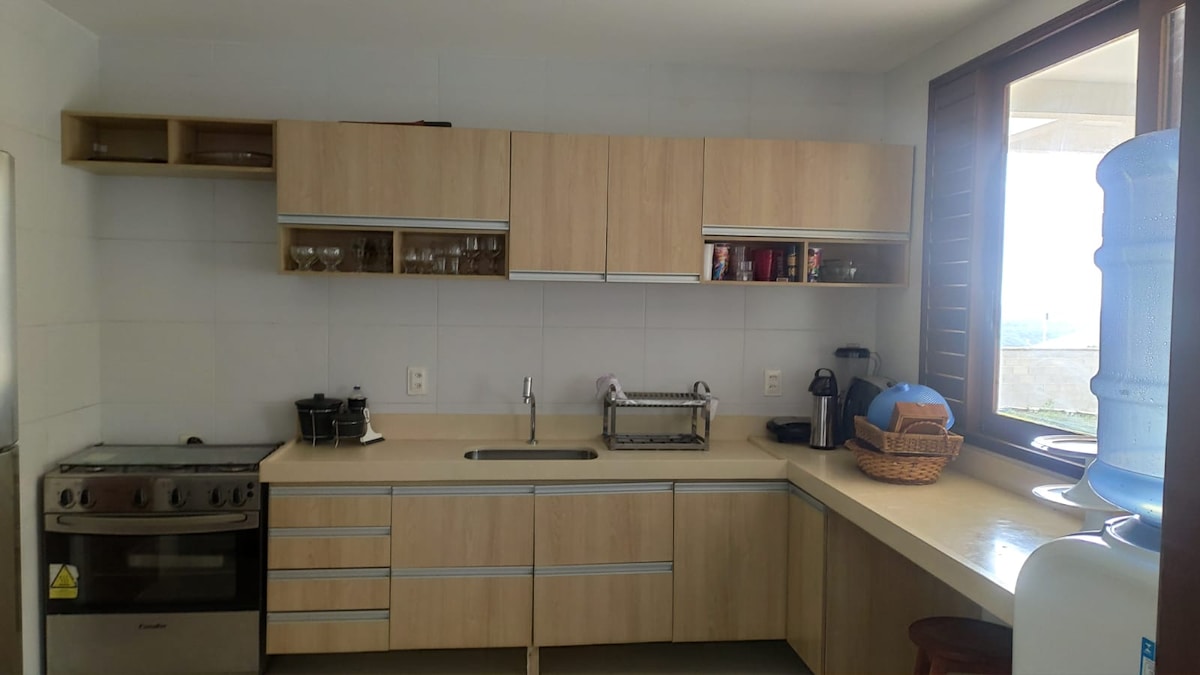 A modern kitchen features light wood cabinetry and a countertop. An oven and stovetop are visible along with a sink and various kitchen appliances. Glassware is neatly arranged on shelves above, and a basket holds kitchen essentials. Natural light filters in through a nearby window.