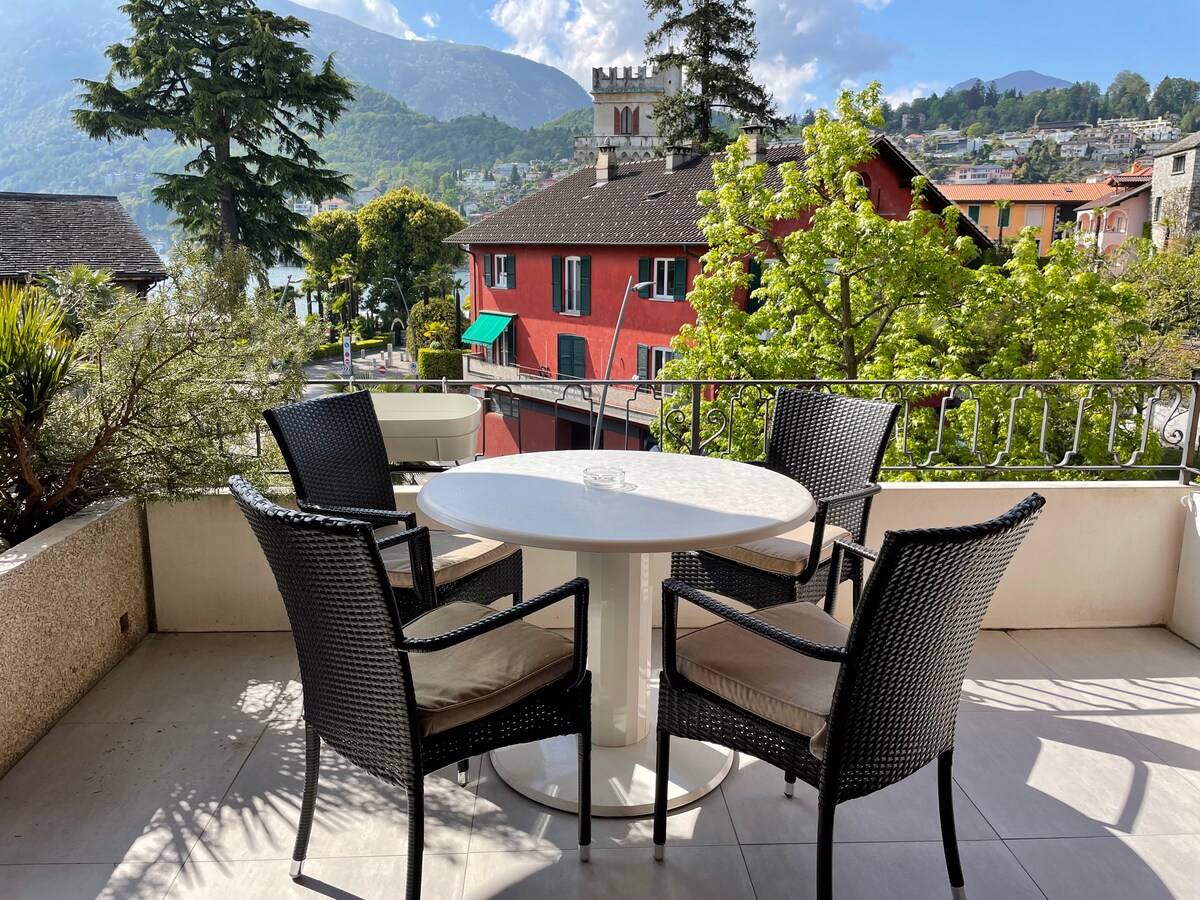 An overed balcony features a round white table surrounded by four black wicker chairs. Lush greenery is visible in the background, along with colorful buildings and mountains in the distance, contributing to a pleasant outdoor space.