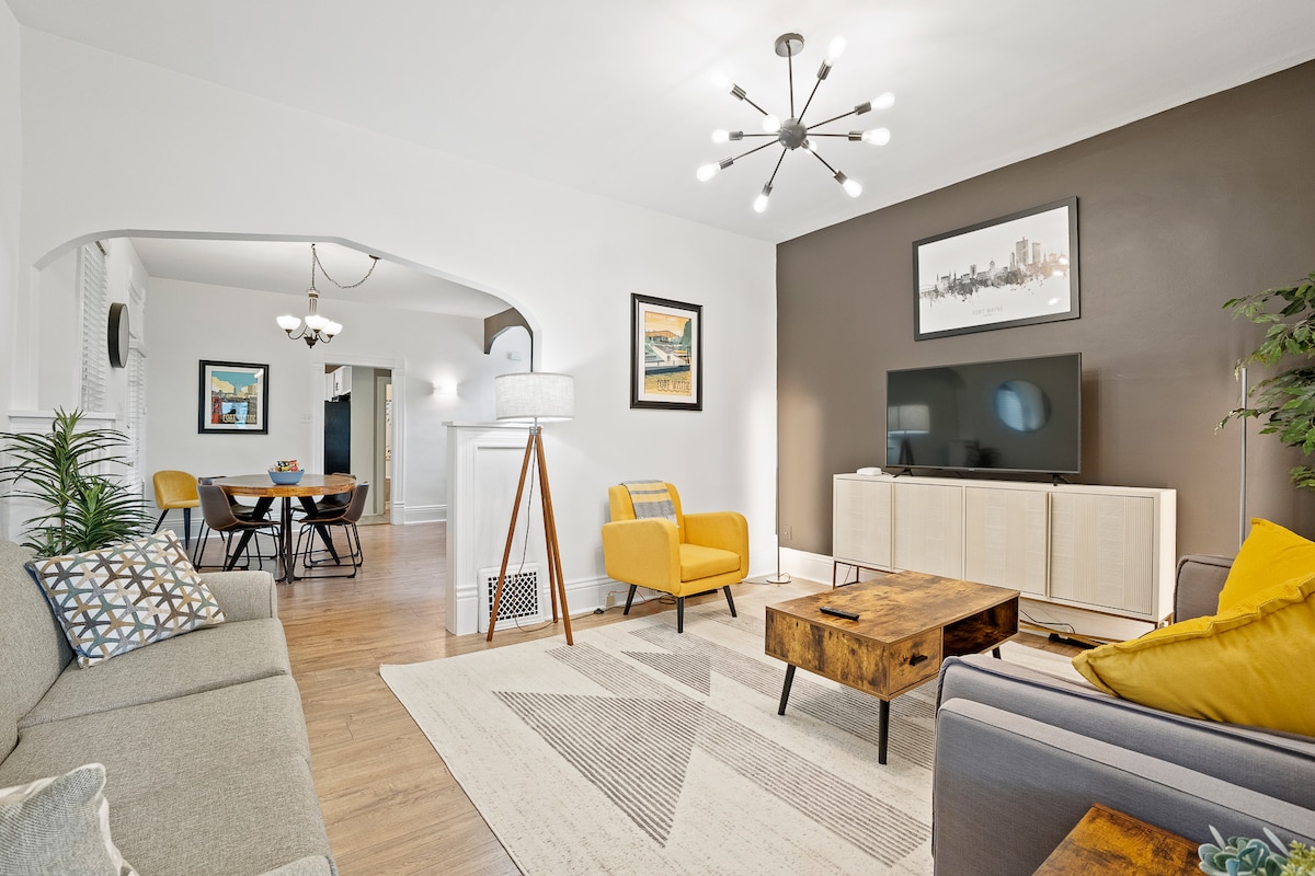 The living room features modern furnishings, including a gray sofa and yellow accent chair, arranged around a wooden coffee table. A smart TV is placed against a dark accent wall, while artwork adorns the walls. The space is illuminated by a contemporary light fixture.