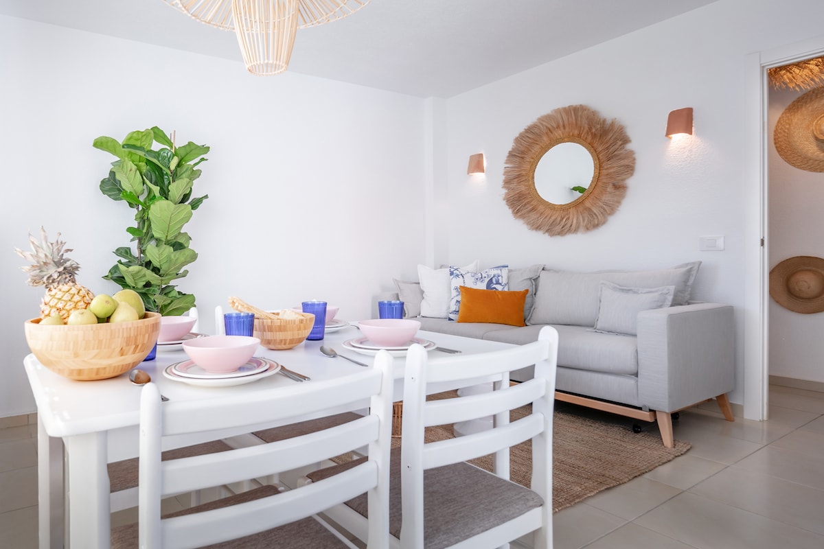 A light and airy living space is showcased, featuring a cozy gray sofa with decorative pillows and a circular mirror on the wall. A dining table is set with colorful tableware, surrounded by white chairs. Lush greenery adds a touch of nature to the room.