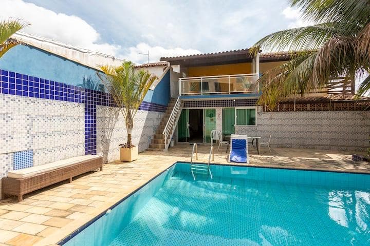 An outdoor area featuring a private pool surrounded by a textured stone deck. Palm trees add greenery around the inviting space. A light brown bench is positioned beside the pool, and a set of stairs leads to the second floor of the house.