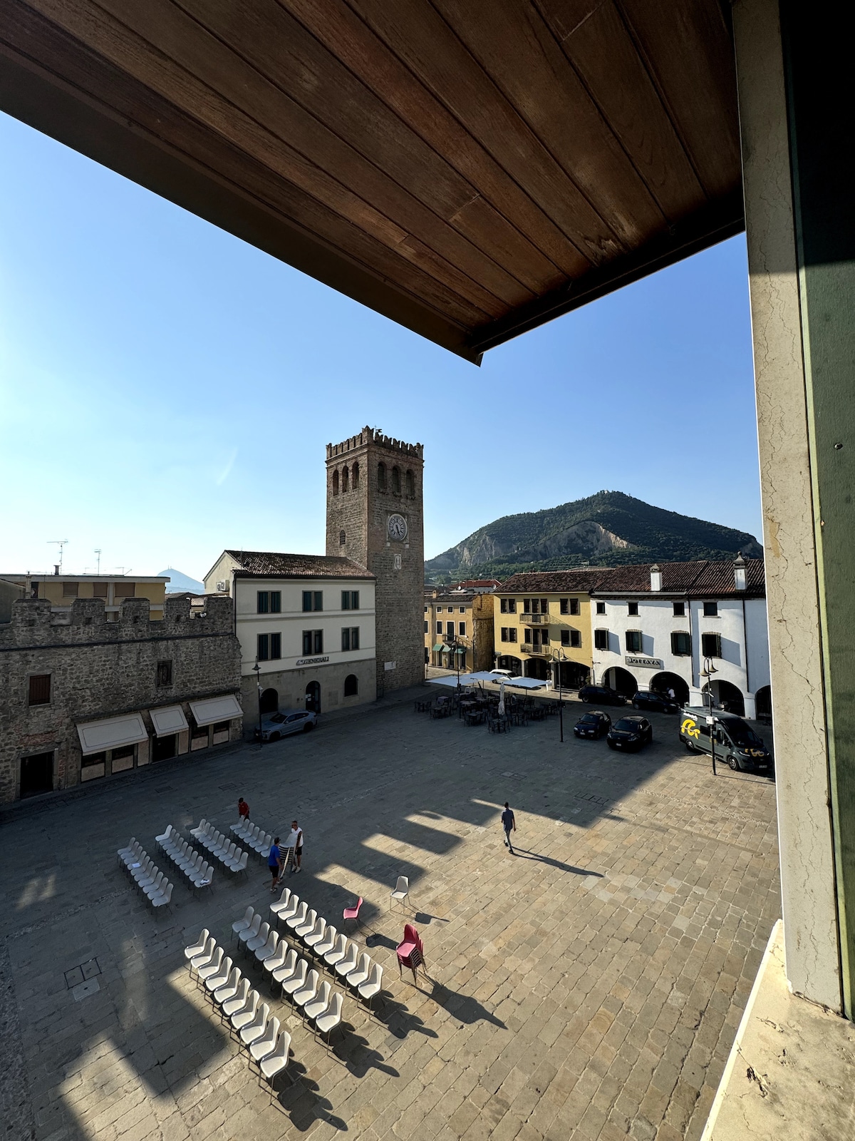 A view of the historic square features a prominent clock tower and surrounding buildings with a variety of architectural styles. Rows of chairs are set up on the cobblestone ground, while people are seen walking. The backdrop includes a mountain under a clear sky.