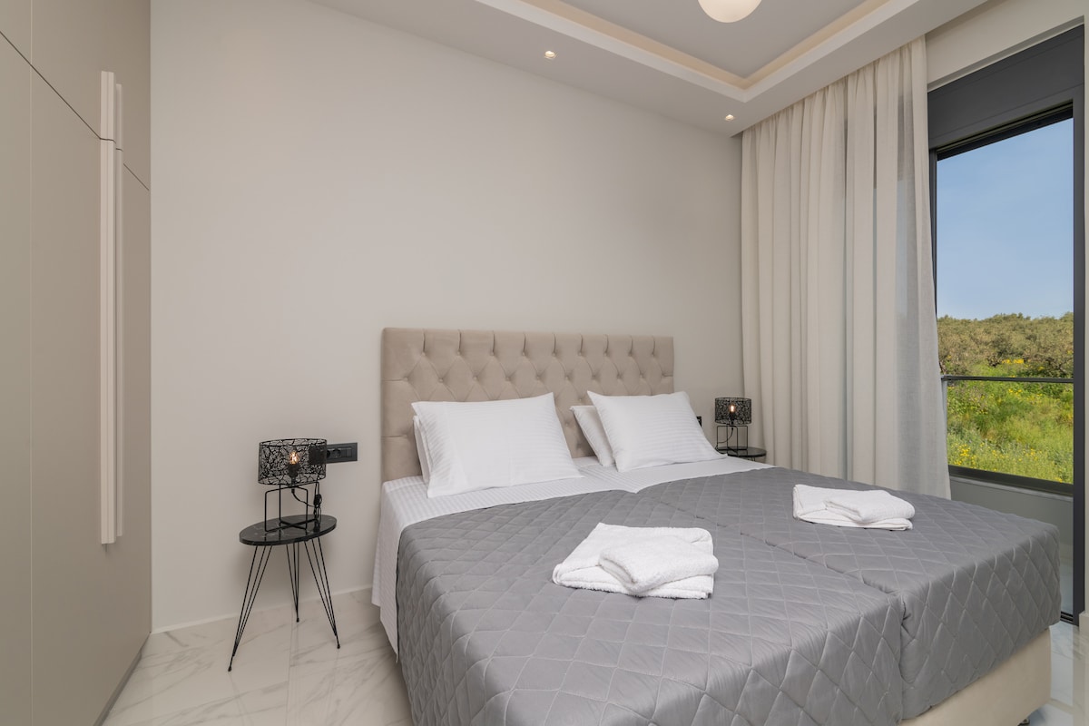 A well-lit bedroom features a spacious bed with a soft headboard, flanked by two bedside tables holding modern lamps. Light curtains frame the window, providing a view of the greenery outside, while fresh towels are neatly arranged on the bed.