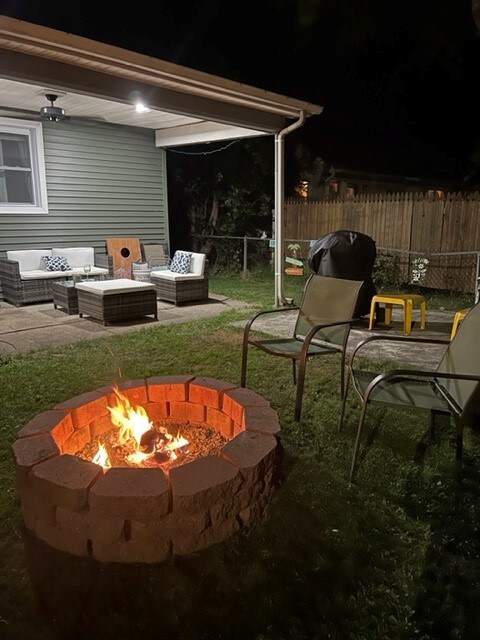 An outdoor space is illuminated by a crackling fire in a stone fire pit, surrounded by metal chairs and a small yellow table. Comfortable seating is arranged nearby under a covered patio, with a view of the lush green yard enclosed by a wooden fence.