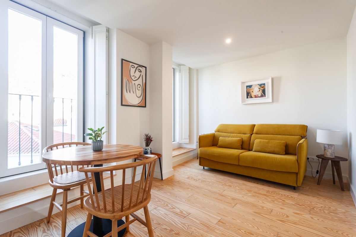 A bright living area features a comfortable yellow sofa and a wooden dining table with two matching chairs. Natural light floods the room through large floor-to-ceiling windows, illuminating a piece of artwork on the wall and a small potted plant atop the table.
