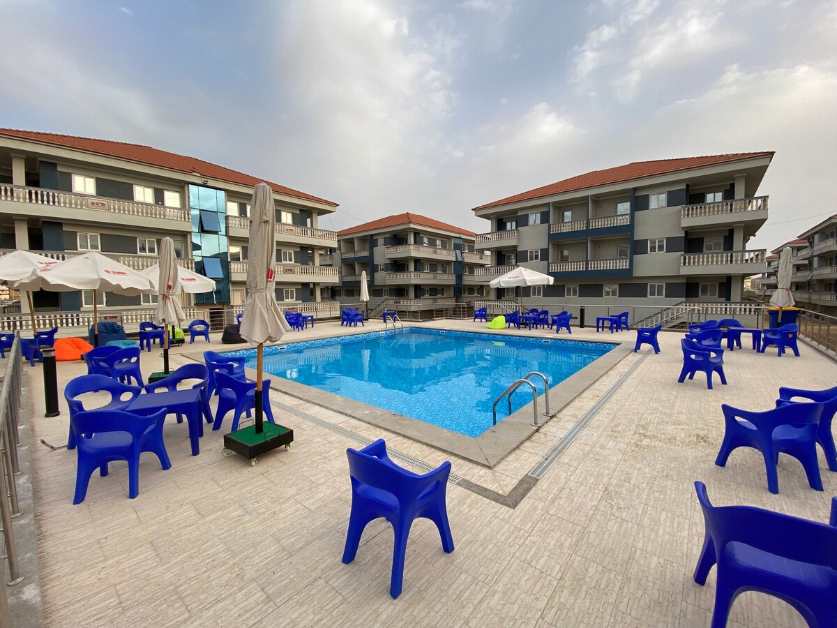 A swimming pool surrounded by a spacious deck features multiple blue chairs arranged around it. Umbrellas provide shade for guests, while modern apartment buildings are visible in the background. The area is designed for relaxation and enjoyment.