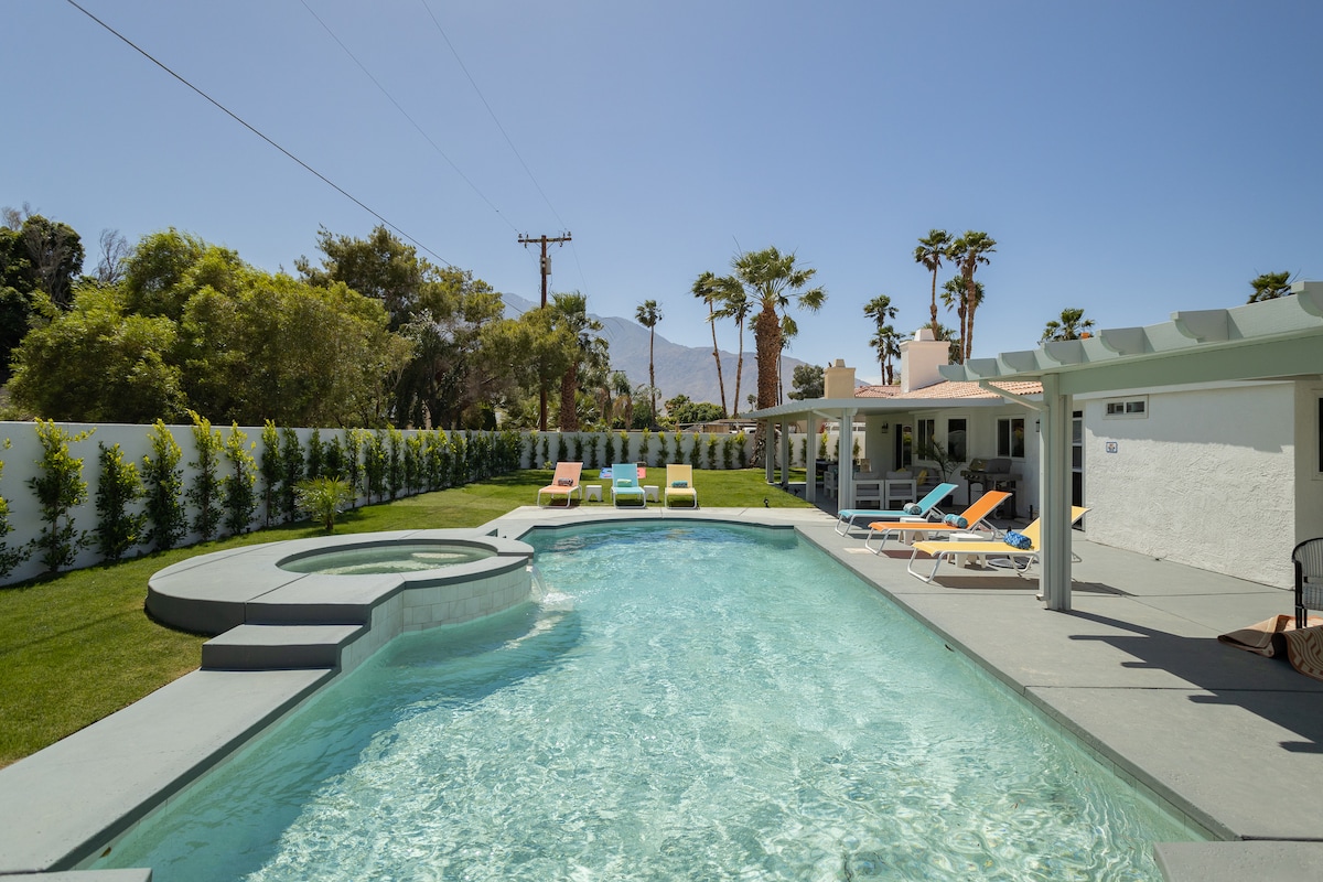 A private backyard features a saltwater pool surrounded by green grass and lounge chairs. A built-in spa is situated next to the pool. Palm trees provide shade, while distant mountains create a scenic backdrop under a clear blue sky.