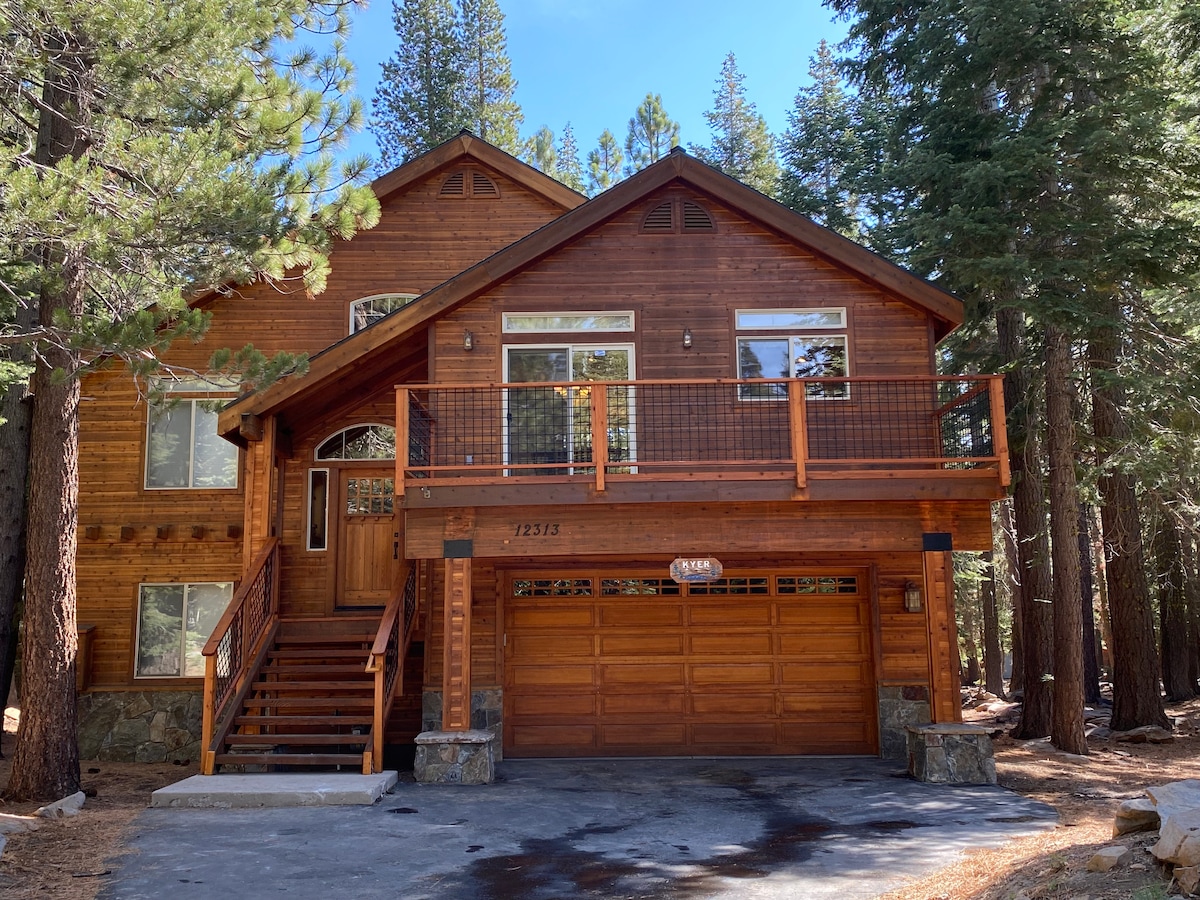 A charming wooden exterior is highlighted by large windows and a welcoming front porch. The home features a two-car garage, complemented by a landscaped area with tall trees offering shade. The structure is set against a natural backdrop of pine trees.