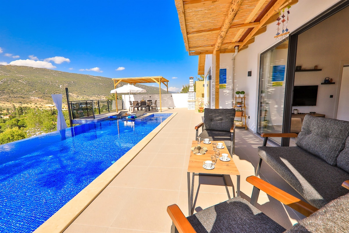 A spacious outdoor terrace features comfortable seating arrangements beside an expansive pool. The clear blue water reflects the sky. A dining area is visible under a pergola, surrounded by natural scenery, creating a serene outdoor environment ideal for relaxation.