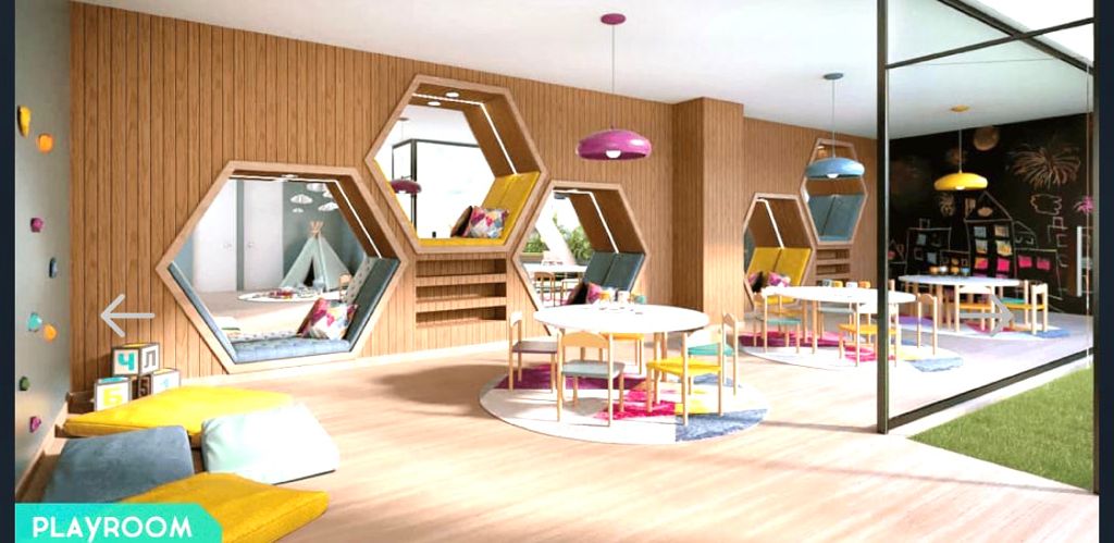 The playroom is bright and spacious, featuring hexagonal mirrors on wooden walls. Colorful tables and chairs are arranged around several play areas, complemented by soft seating in yellow and blue. Large windows allow natural light to fill the room, creating a cheerful environment for children.