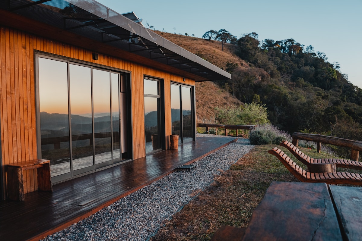 The exterior showcases a modern wooden cabin with large glass doors reflecting the surrounding landscape. A spacious deck features comfortable wooden loungers and a dining table. The backdrop includes rolling hills and natural vegetation, creating a serene outdoor setting.