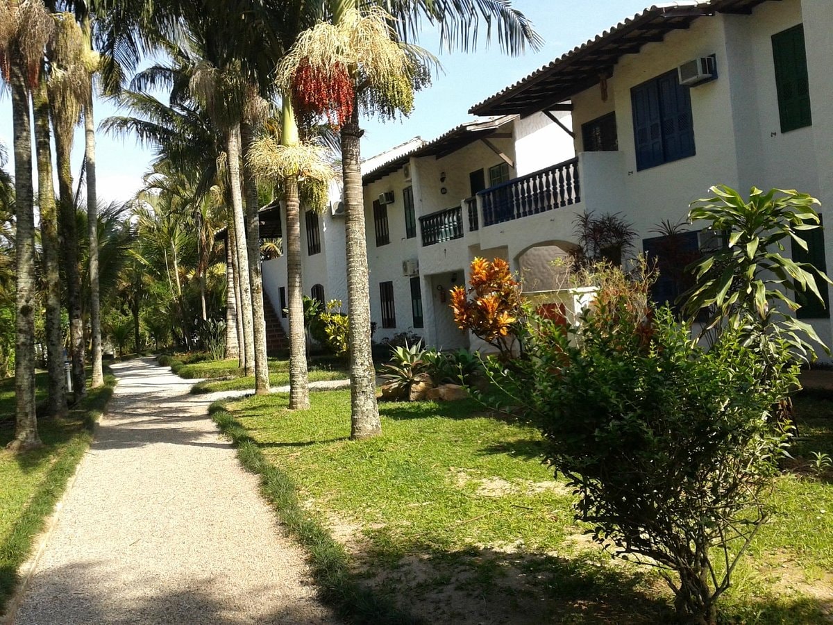A scenic pathway meanders through lush greenery alongside two-story buildings adorned with blue-green shutters. Tropical palm trees provide shade, while colorful plants add visual interest. The surroundings suggest a tranquil atmosphere, inviting guests to explore the natural beauty of the resort.