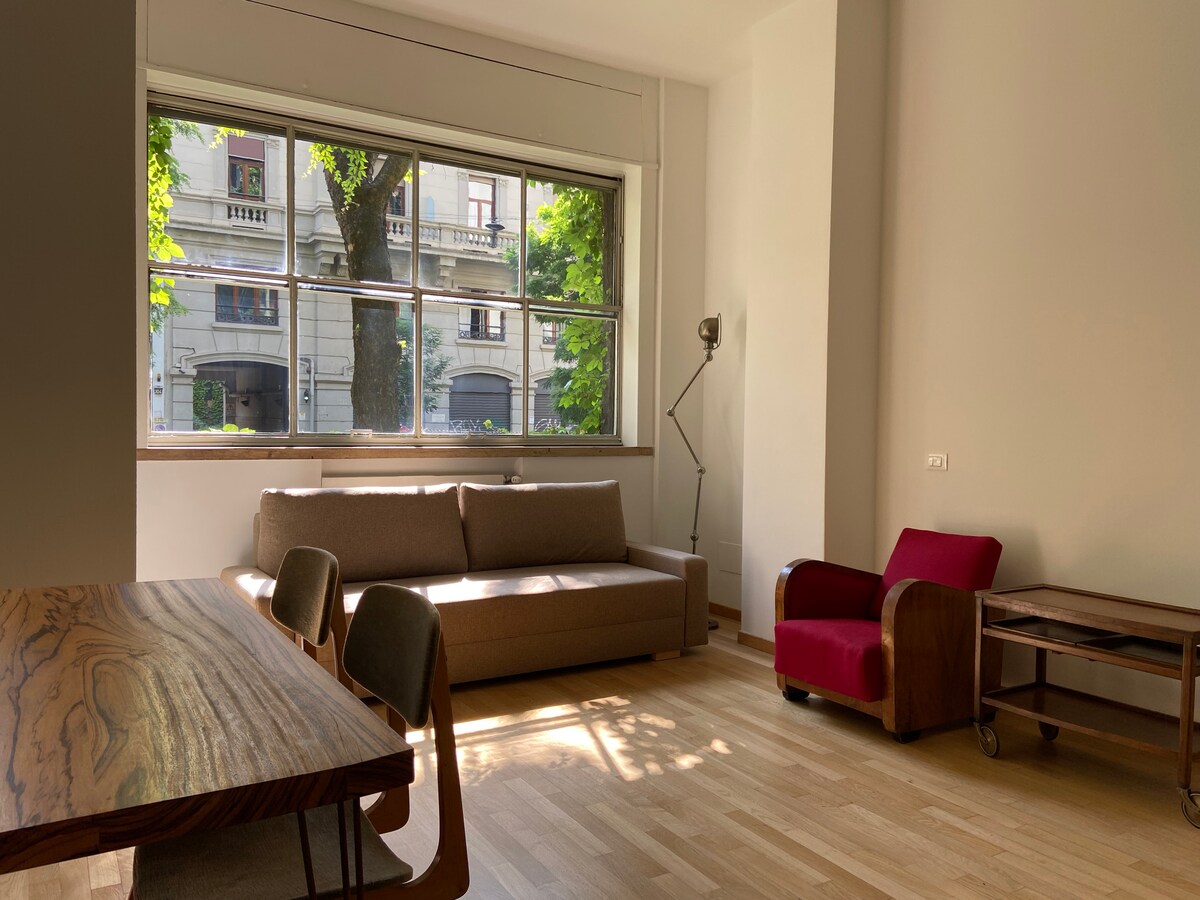 A bright living area features a large window overlooking a garden, allowing ample natural light to enter. A spacious sofa and a vintage red armchair are positioned adjacent to a wooden dining table, enhancing the uncluttered design of the open space.