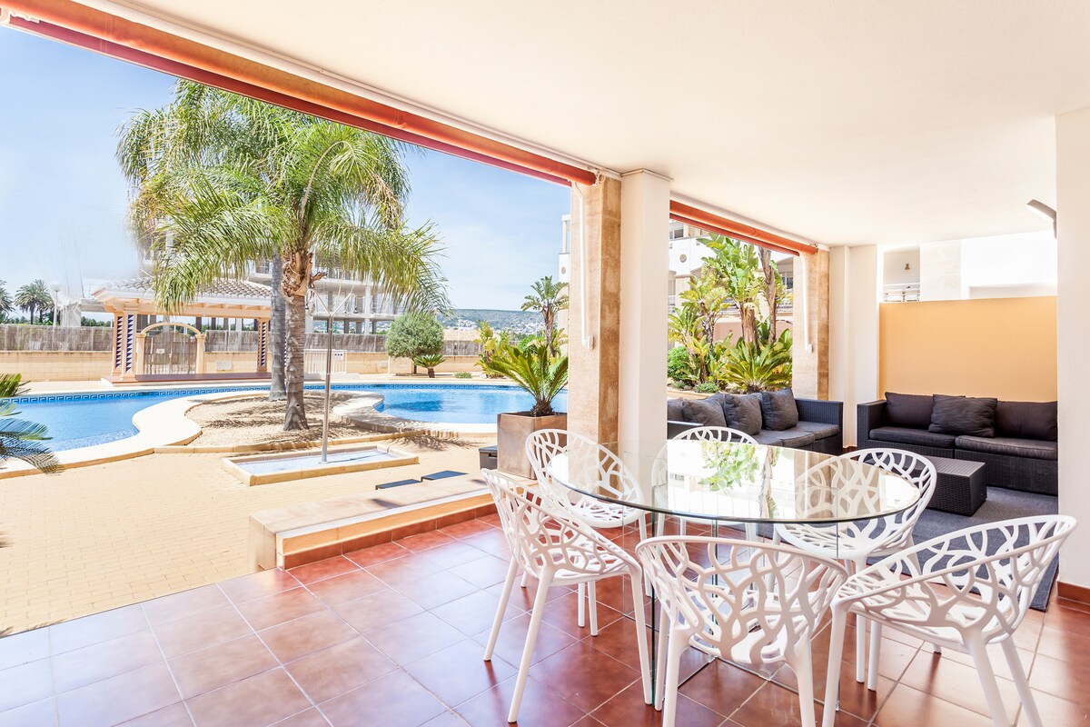 An outdoor seating area features a glass table surrounded by white chairs. Lush greenery and palm trees frame the scene, with a large pool visible in the background. Sunlight brightens the space, enhancing the open atmosphere.