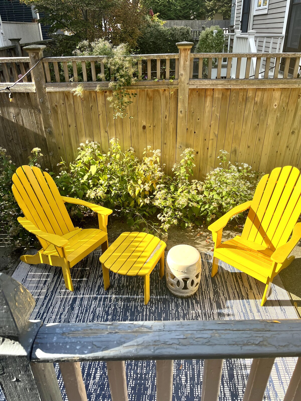 A small outdoor patio features two vibrant yellow adirondack chairs paired with a round side table. The area is surrounded by a wooden fence and greenery, creating a cozy and inviting space for relaxation.