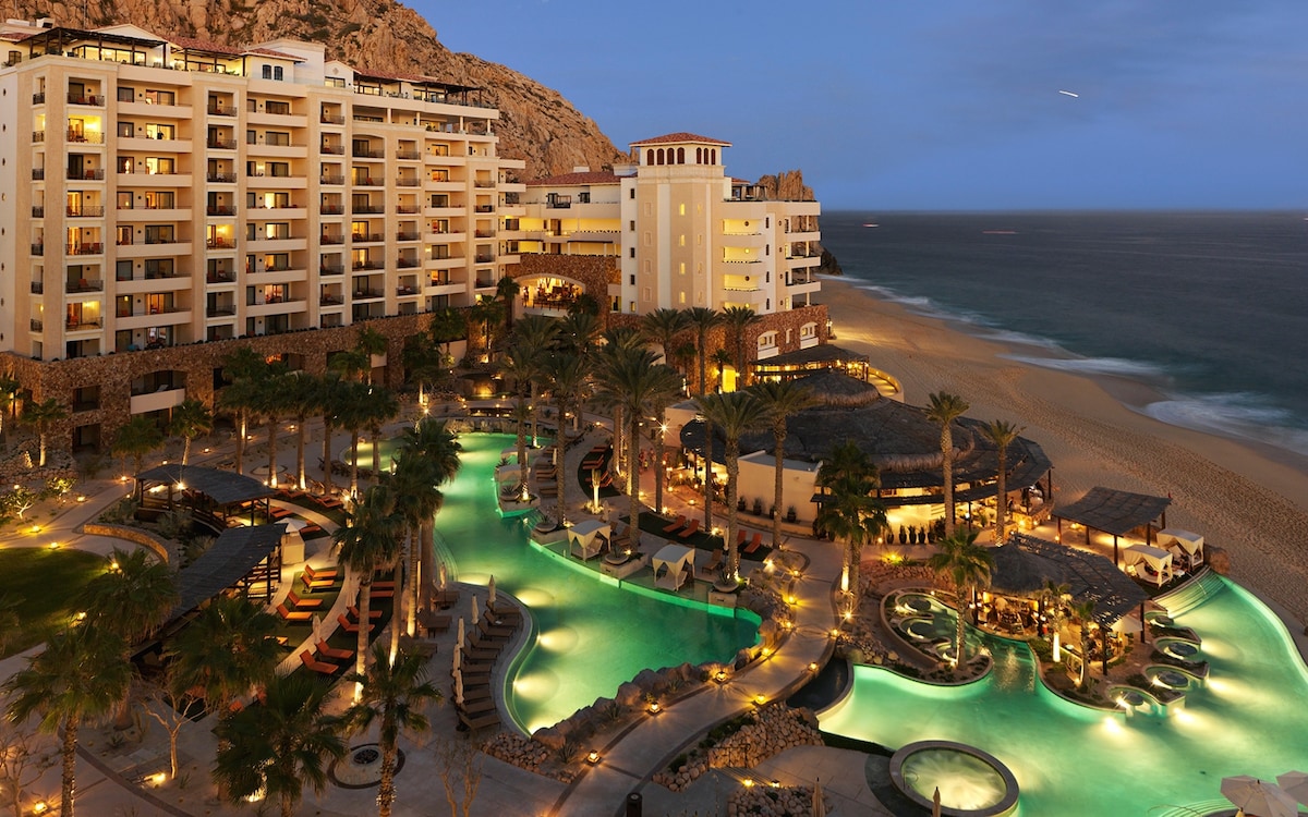 An expansive resort is depicted at twilight, featuring multiple illuminated swimming pools surrounded by palm trees. Beachside restaurants and lounge areas are visible, with waves gently lapping at the shore in the background. The building's architecture blends modern design with natural elements.