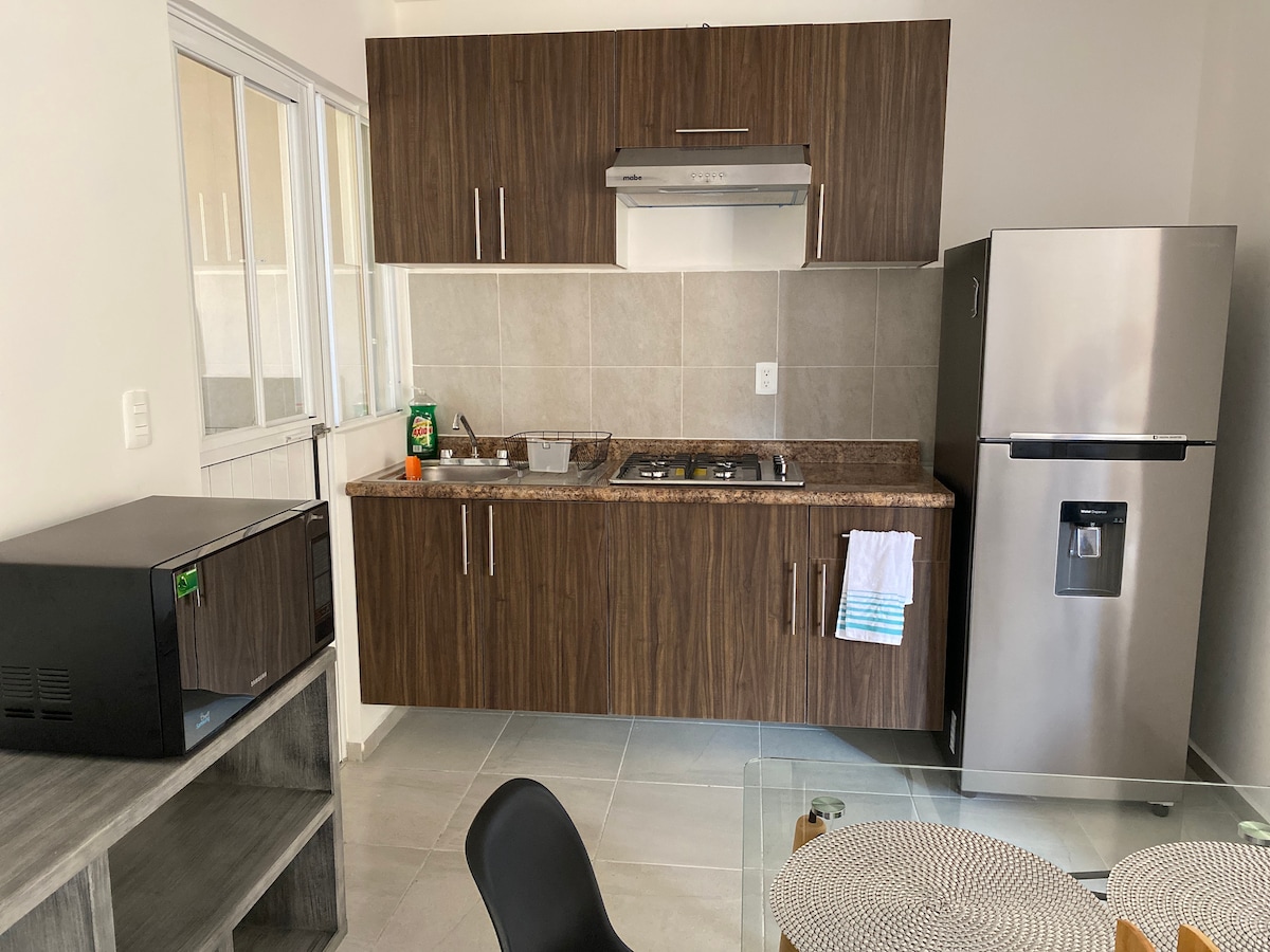 A modern kitchen area is presented with wooden cabinets and a granite countertop. A stove with an exhaust hood and a microwave are located alongside a refrigerator. Natural light enters through the nearby windows, highlighting the organized layout and essential appliances.