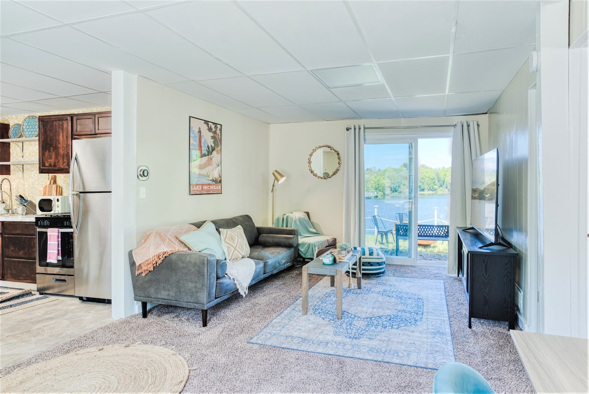 A living room with a plush sofa faces a coffee table, complemented by a light area rug. A large sliding door provides access to a patio with lake views. Soft natural light fills the space, and a small dining area is visible in the background.