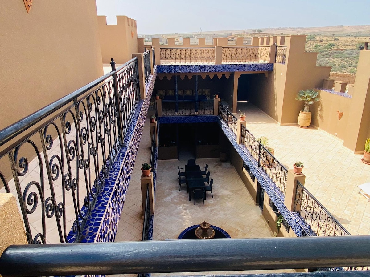 A spacious courtyard is framed by ornate wrought iron railings and features terracotta planters. A seating arrangement of black chairs surrounds a low table at the center, with a decorative fountain adding an element of tranquility. Surrounding walls showcase traditional Moroccan blue tile accents.