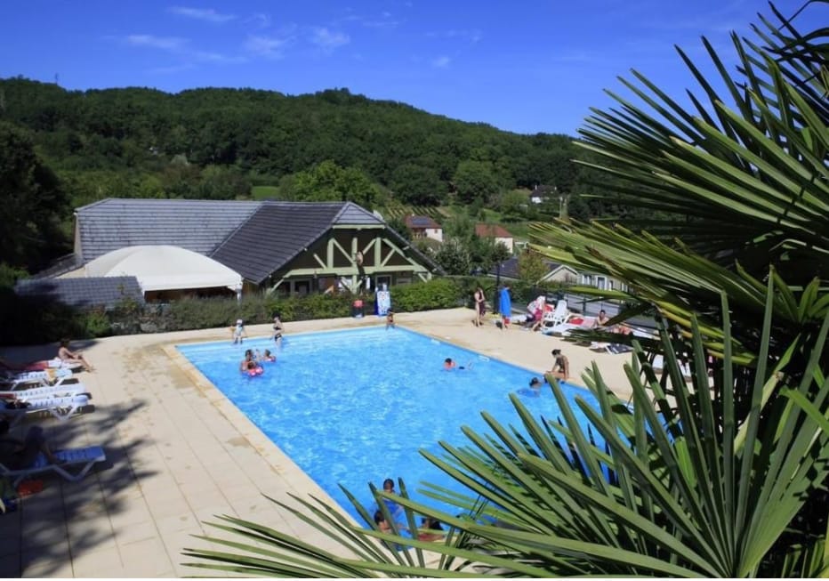 A large outdoor swimming pool is surrounded by a sun patio with lounge chairs. Families can be seen enjoying the water, while lush green hills rise in the background, complemented by a charming building with a sloped roof nearby.