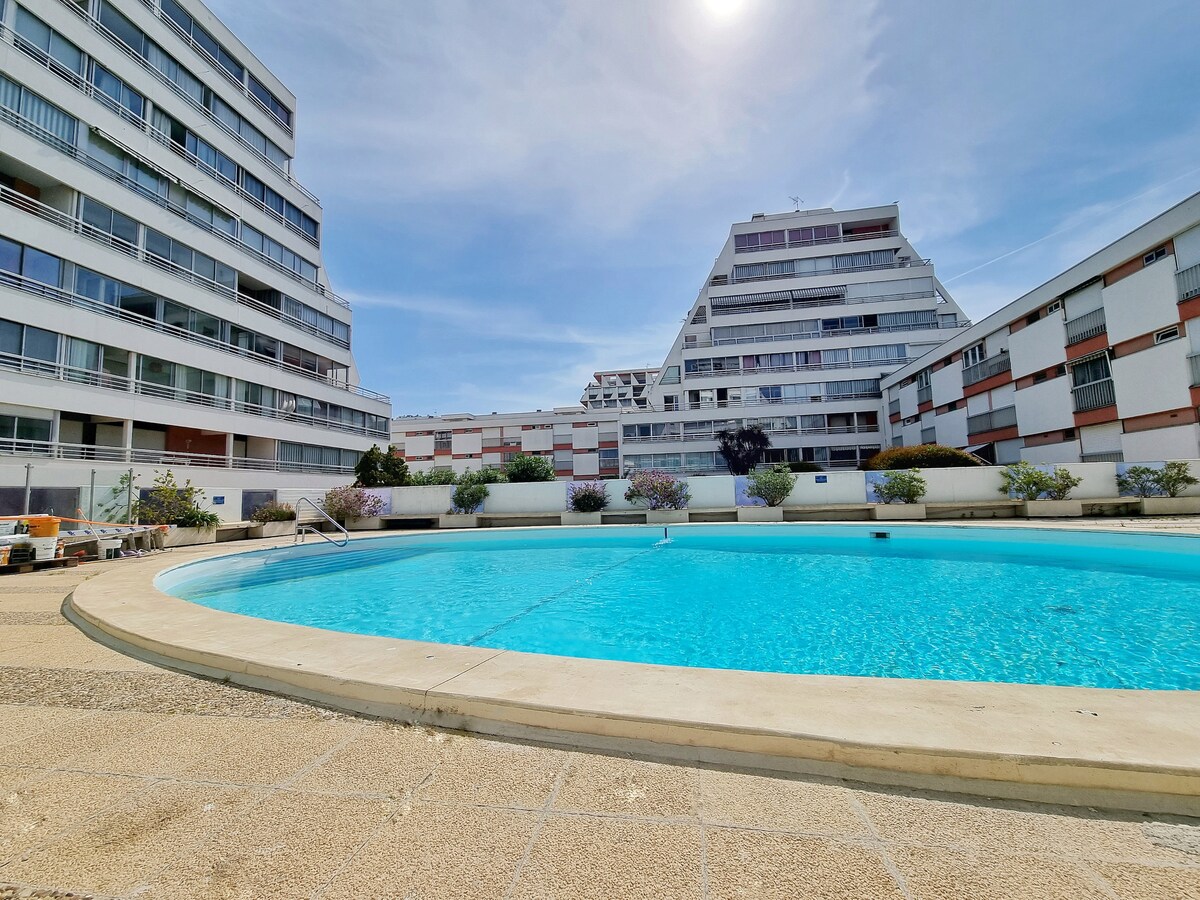 A private swimming pool is surrounded by landscaped areas, featuring low shrubbery and sunlit concrete. The pool's water reflects the clear blue sky above and is accessible from the adjacent buildings, which are designed with multiple balconies and modern architecture.