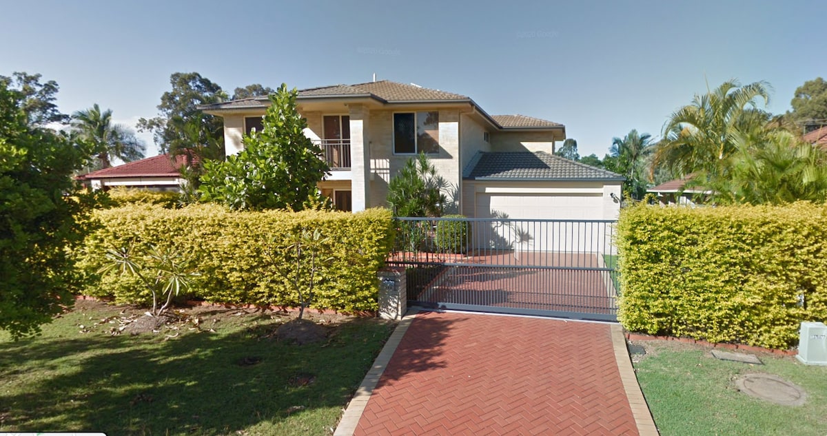 A two-story home is shown, featuring a beige exterior with a tile roof. The entrance is framed by neatly trimmed hedges and vibrant greenery, with a gated driveway leading to the front door. Palm trees and shrubs add to the landscape.