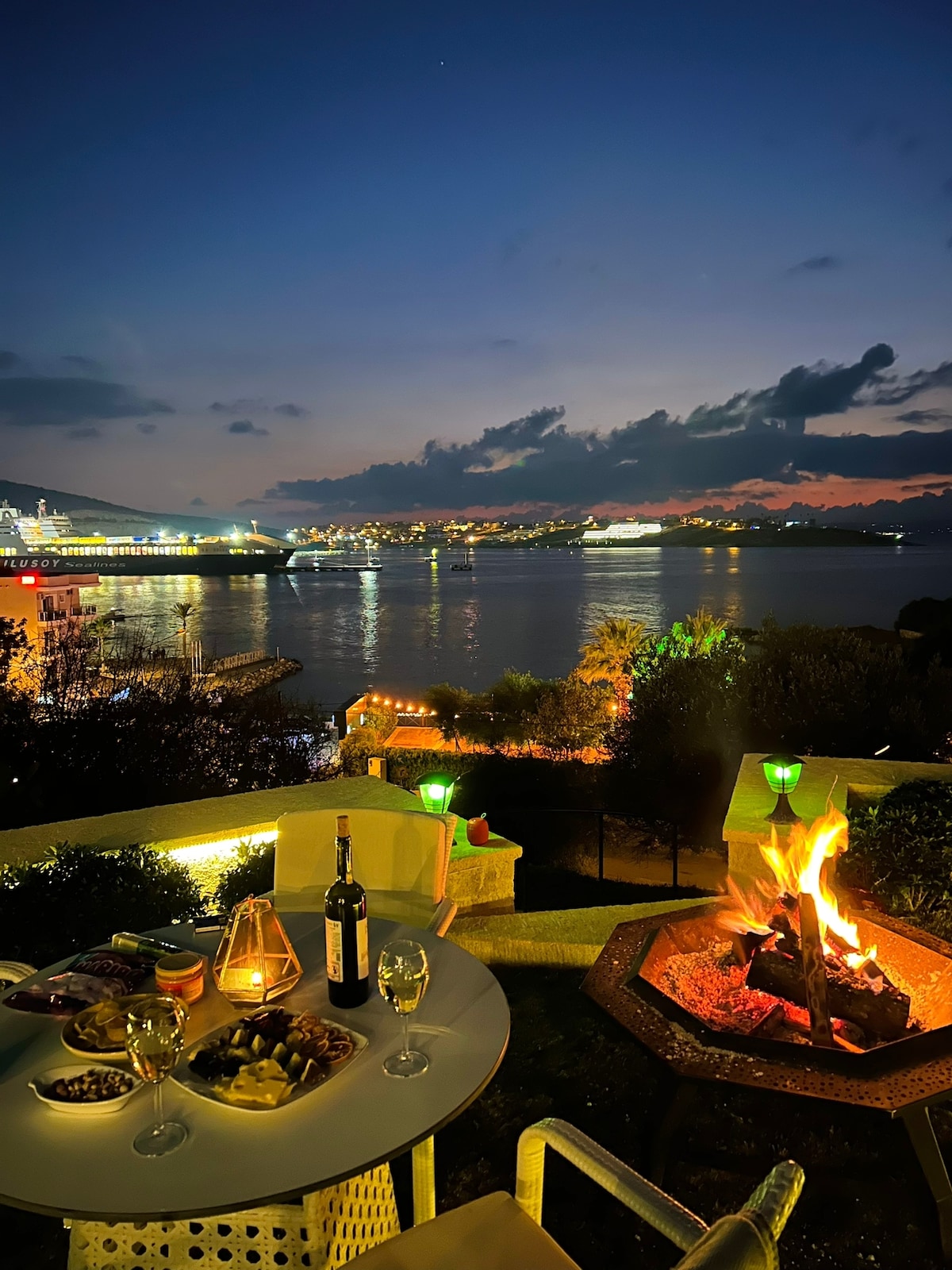An outdoor seating area is displayed with a table set for two, featuring glasses of wine and an assortment of snacks. A fire pit is visible, with flames glowing warmly against the backdrop of a scenic waterfront at dusk, illuminated by city lights and a colorful sky.