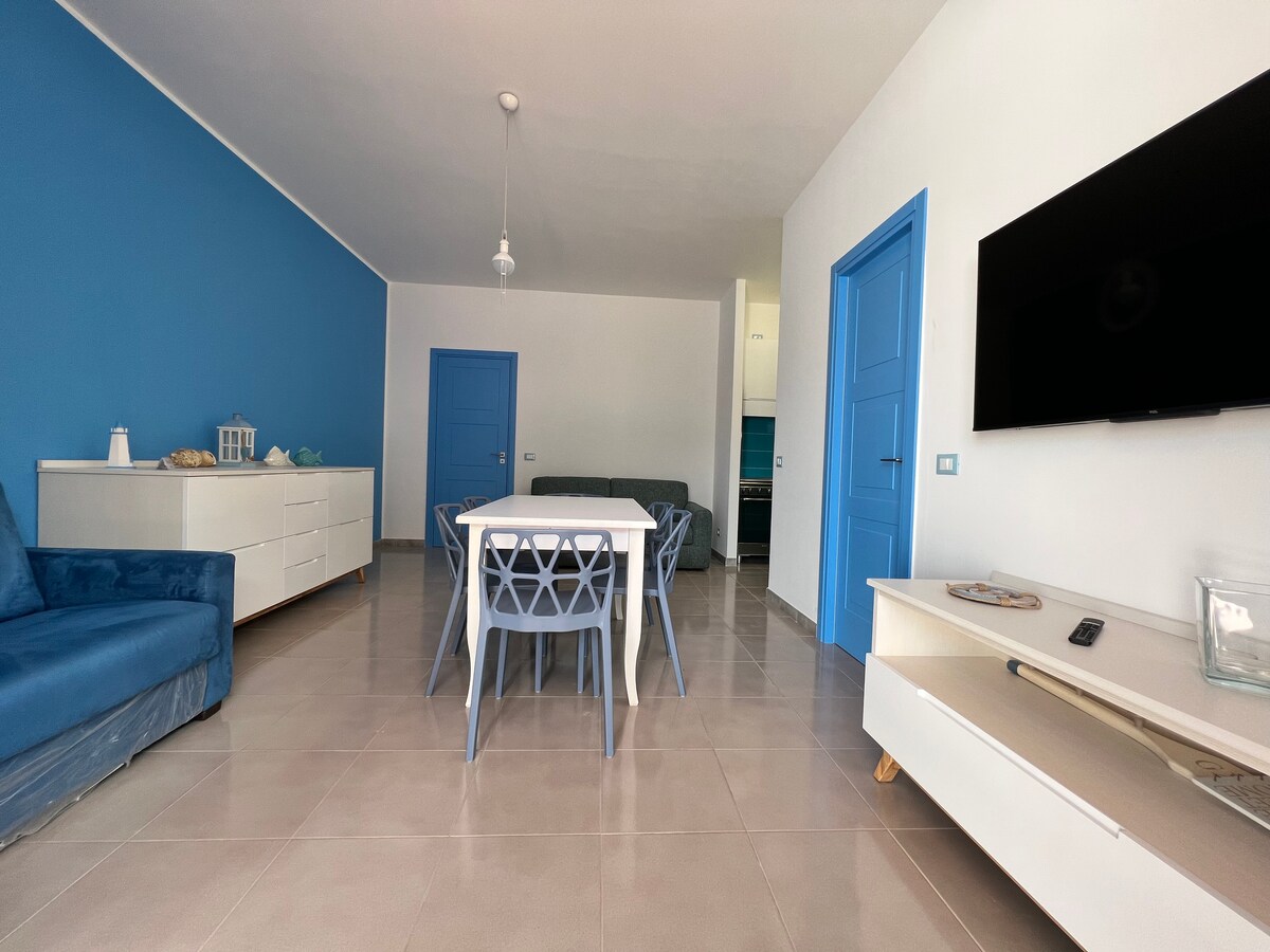 A bright living space features a blue accent wall and modern furnishings. A dining table with light blue chairs is positioned at center. A comfortable blue sofa sits to one side, while a TV is mounted on the wall opposite a sleek entertainment unit.