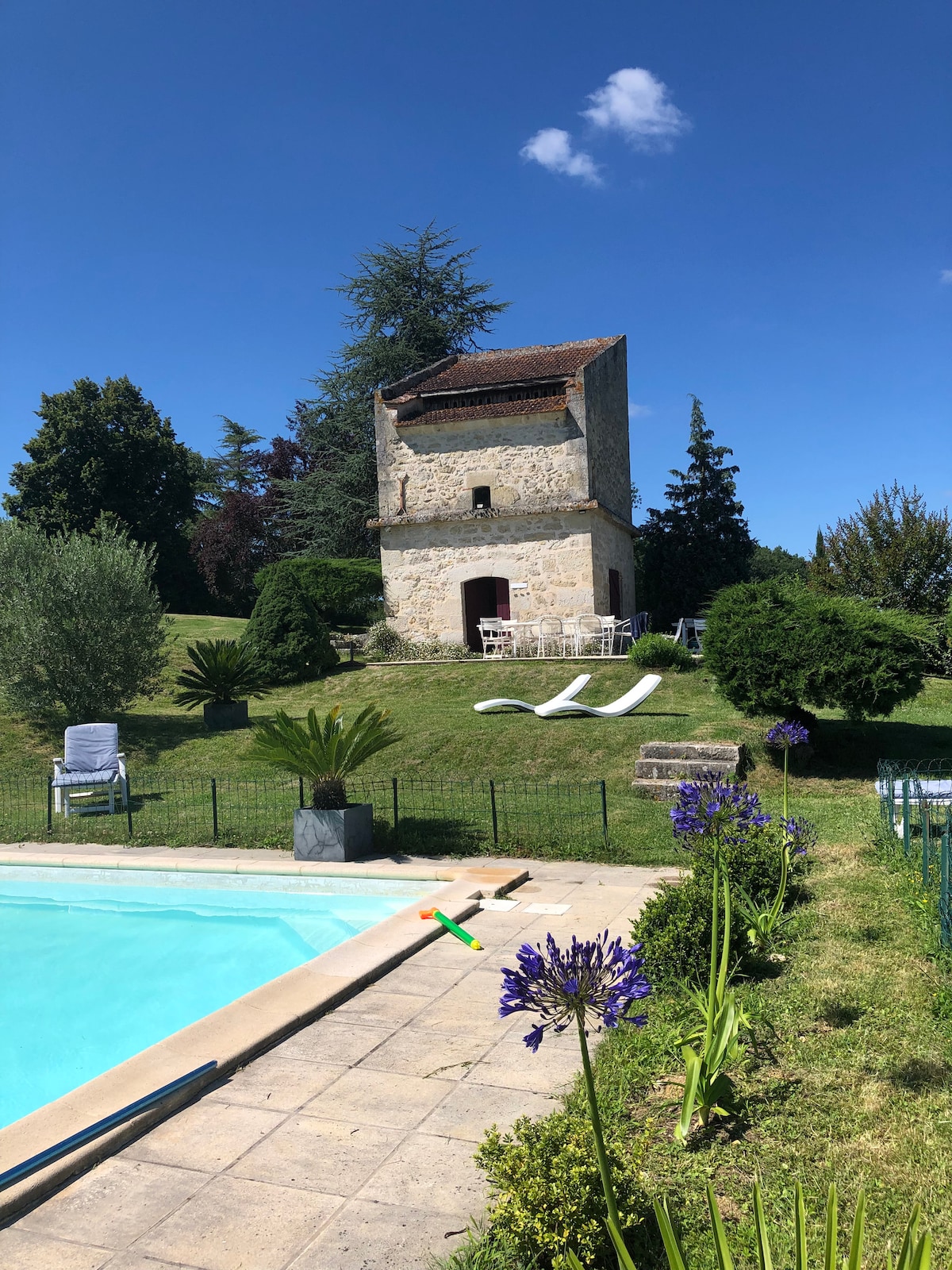 A stone structure is visible on a gently sloping hill, surrounded by lush greenery. A swimming pool is located in the foreground, complemented by a well-maintained garden featuring flowering plants. Outdoor seating is arranged near the pool, creating a serene outdoor space.