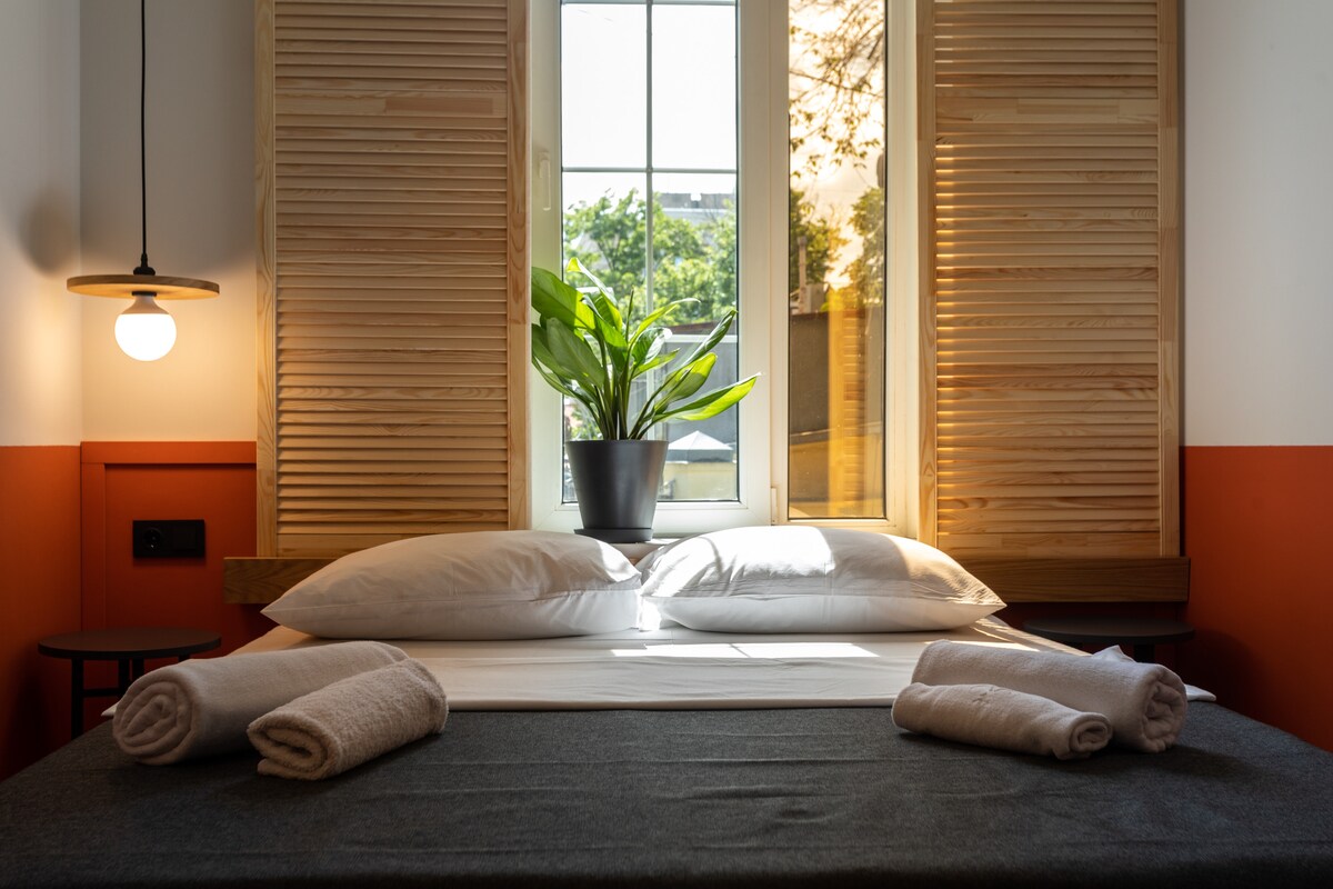A well-lit bedroom features a spacious bed with soft linens and two rolled towels placed neatly at the foot. Natural light enters through a window flanked by wooden shutters, illuminating a potted plant on the nightstand.