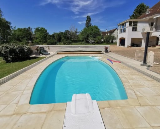 A clear swimming pool is presented, surrounded by a spacious deck. Lush greenery can be seen in the background, while a patio area and building are visible beyond the pool. The inviting turquoise water reflects the blue sky with few clouds.