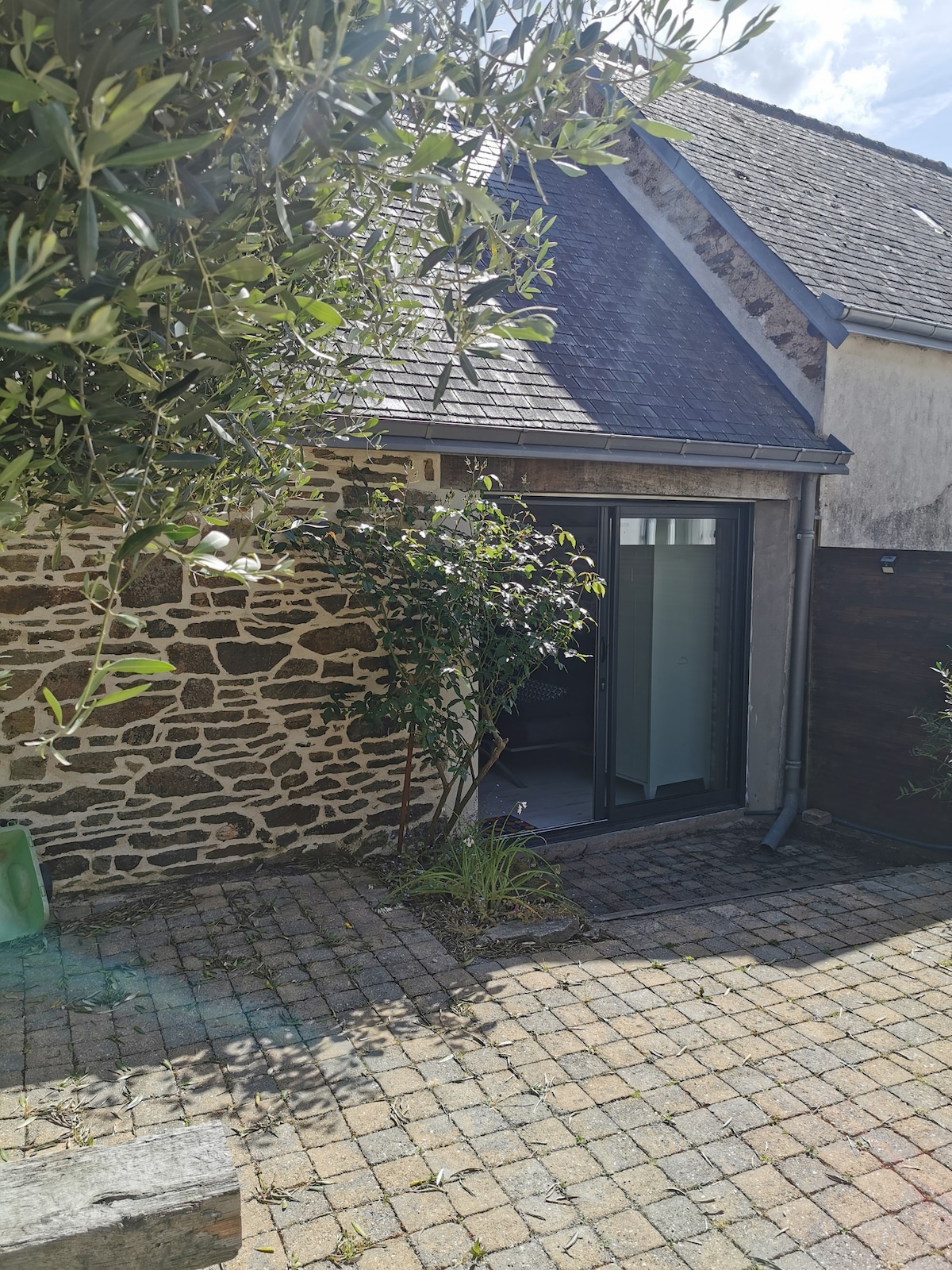 An exterior view of the studio showcases a stone facade complemented by large glass doors that open to a small enclosed courtyard. Surrounding greenery and a stone pathway create a serene and inviting entrance atmosphere.