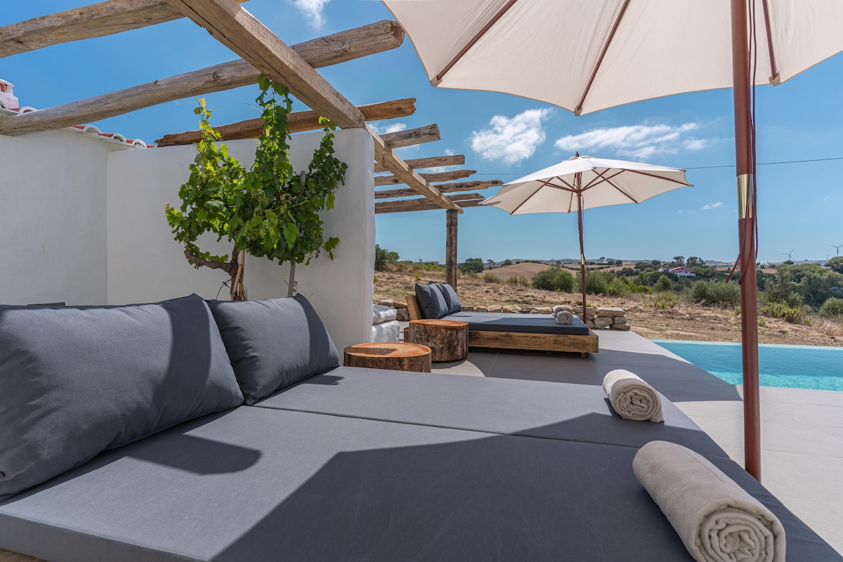 A lounge area is presented with large gray cushions arranged on a spacious deck, accompanied by wooden tables. Two umbrellas offer shade, while a vine grows nearby. In the background, rolling hills and a clear blue sky enhance the tranquil setting.